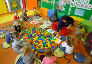 Dzieci siedzą wokół kolorowych piłek i pojemników, pani Ewelina tłumaczy dzieciom w jaki kolor piłek wkładamy do danego pojemnika.